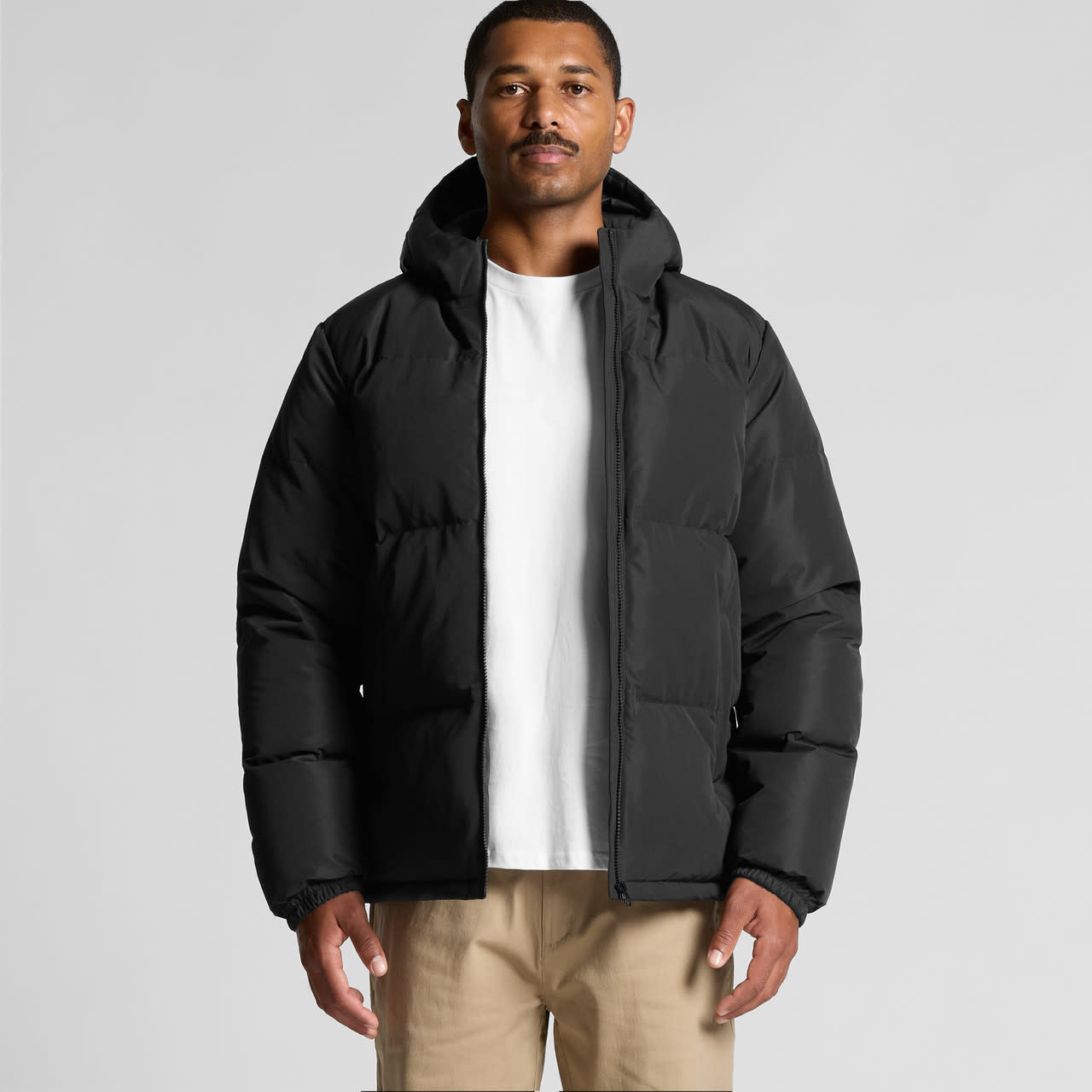 A black hooded puffer jacket featuring a front zipper and a logo, worn by a model against a neutral background. Hooded Puffer Jacket - Image 1