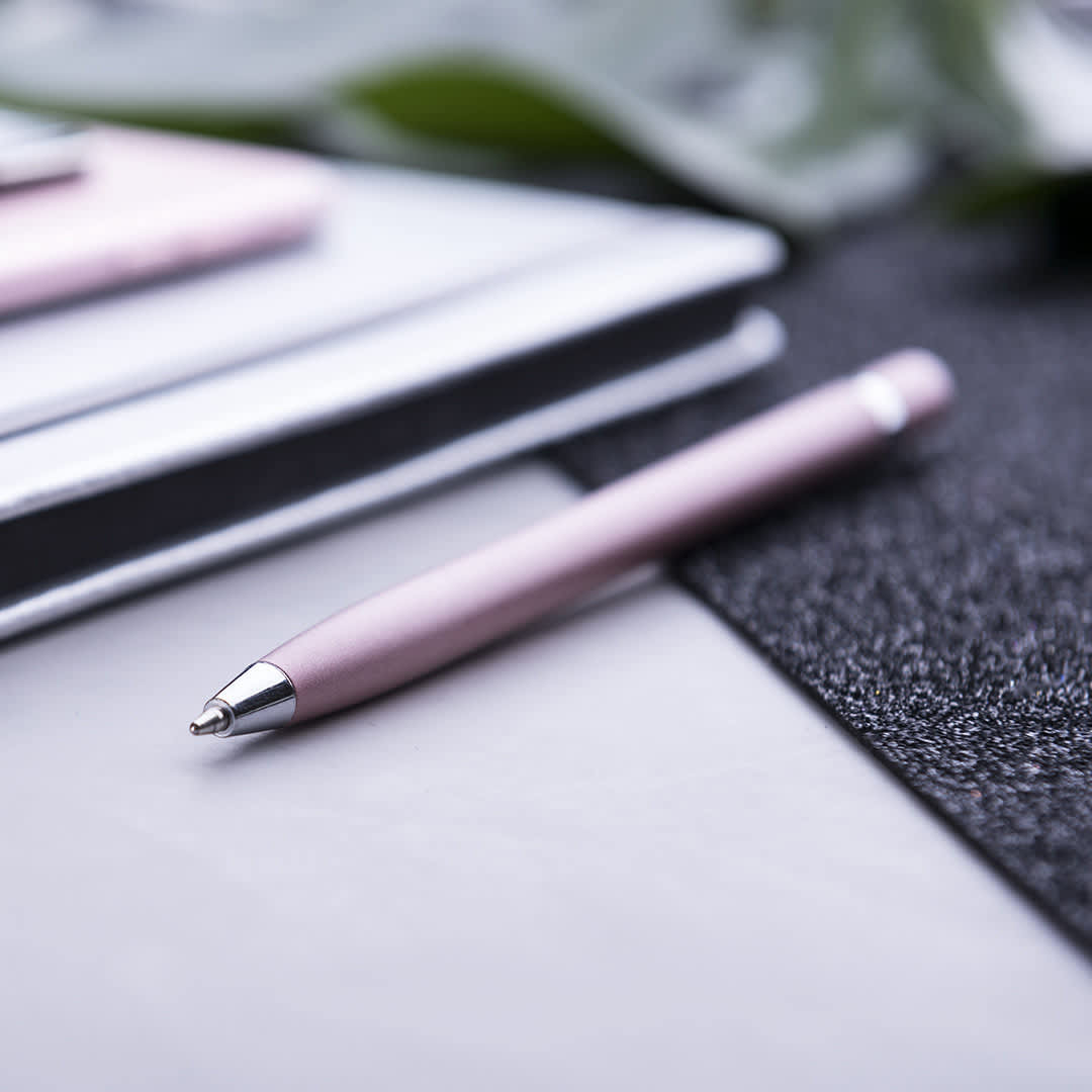 A pink pen with a silver tip rests on a textured surface, alongside a notebook and other stationery. Mulent - Image 1