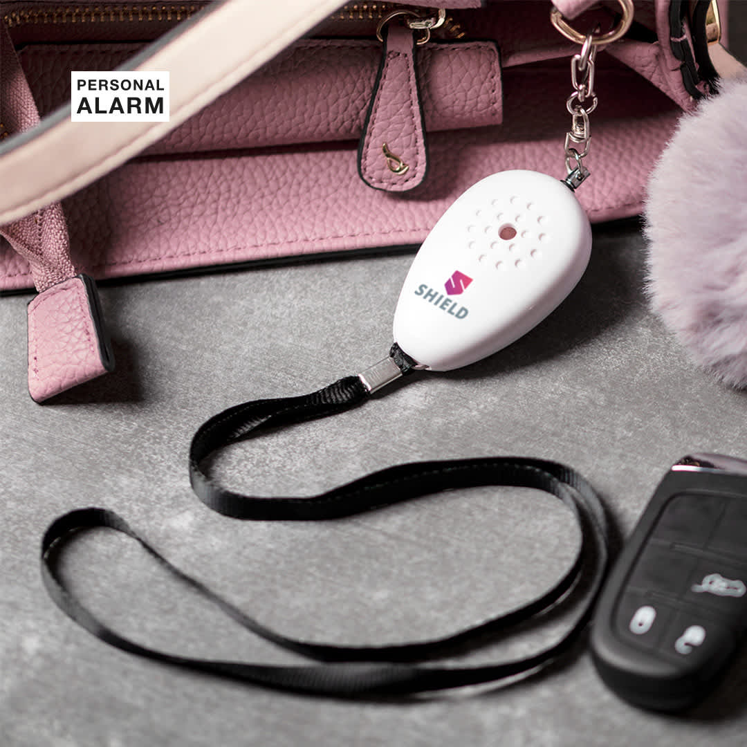 A white personal alarm with a black strap, placed beside a pink handbag and a car key fob. Birnal Personal Alarm - Image 1