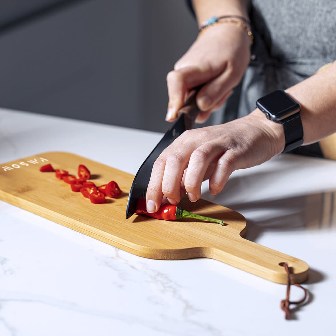 A chopping board made of bamboo with a hand using a black knife to slice red chillies. The board has a brand logo. Saraby - Image 1