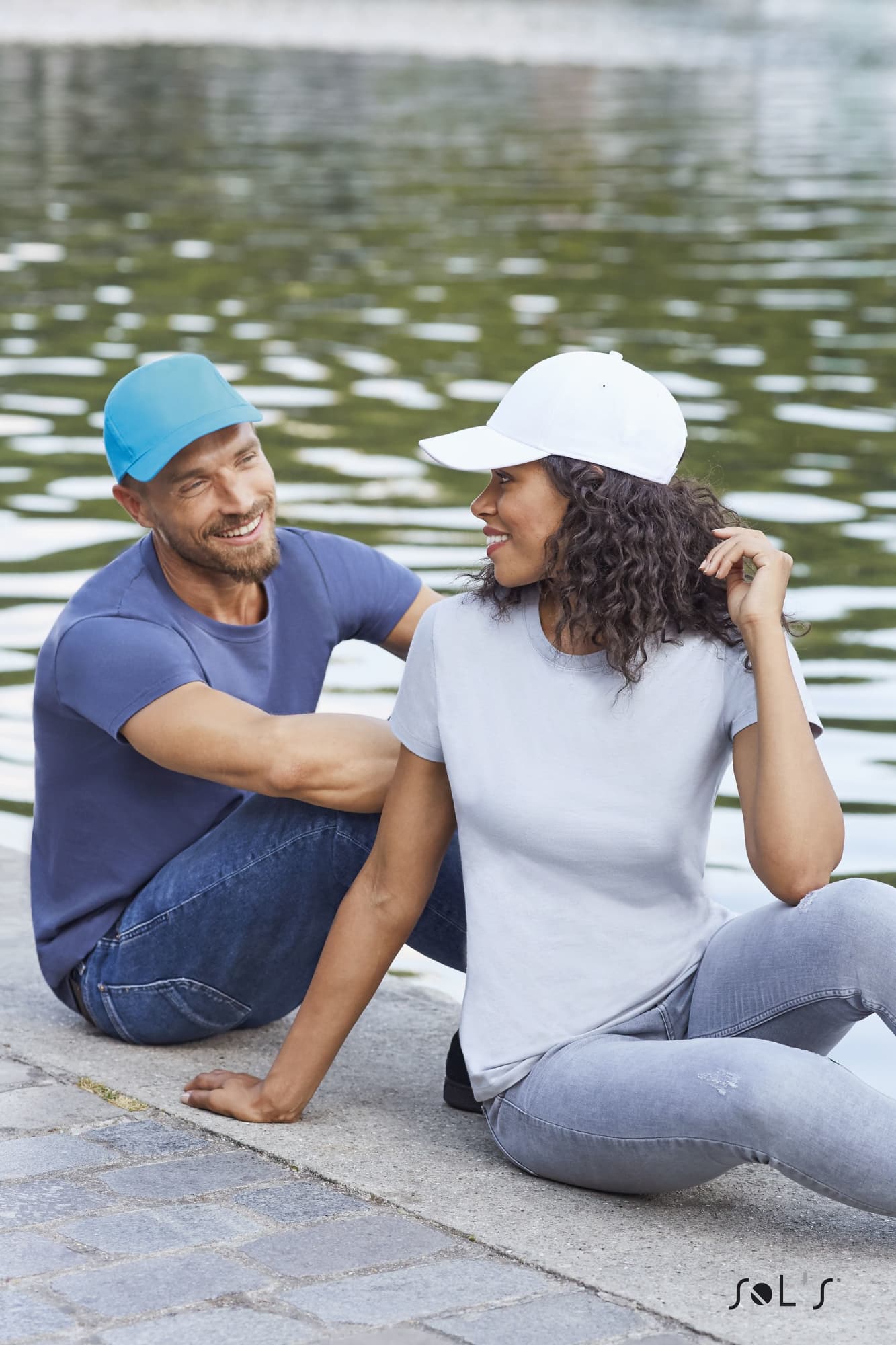 Two models by a water feature, one wearing a light blue cap and the other in a white cap, both in casual attire. Cap 5 panel light brushed cotton SUNNY - Image 1