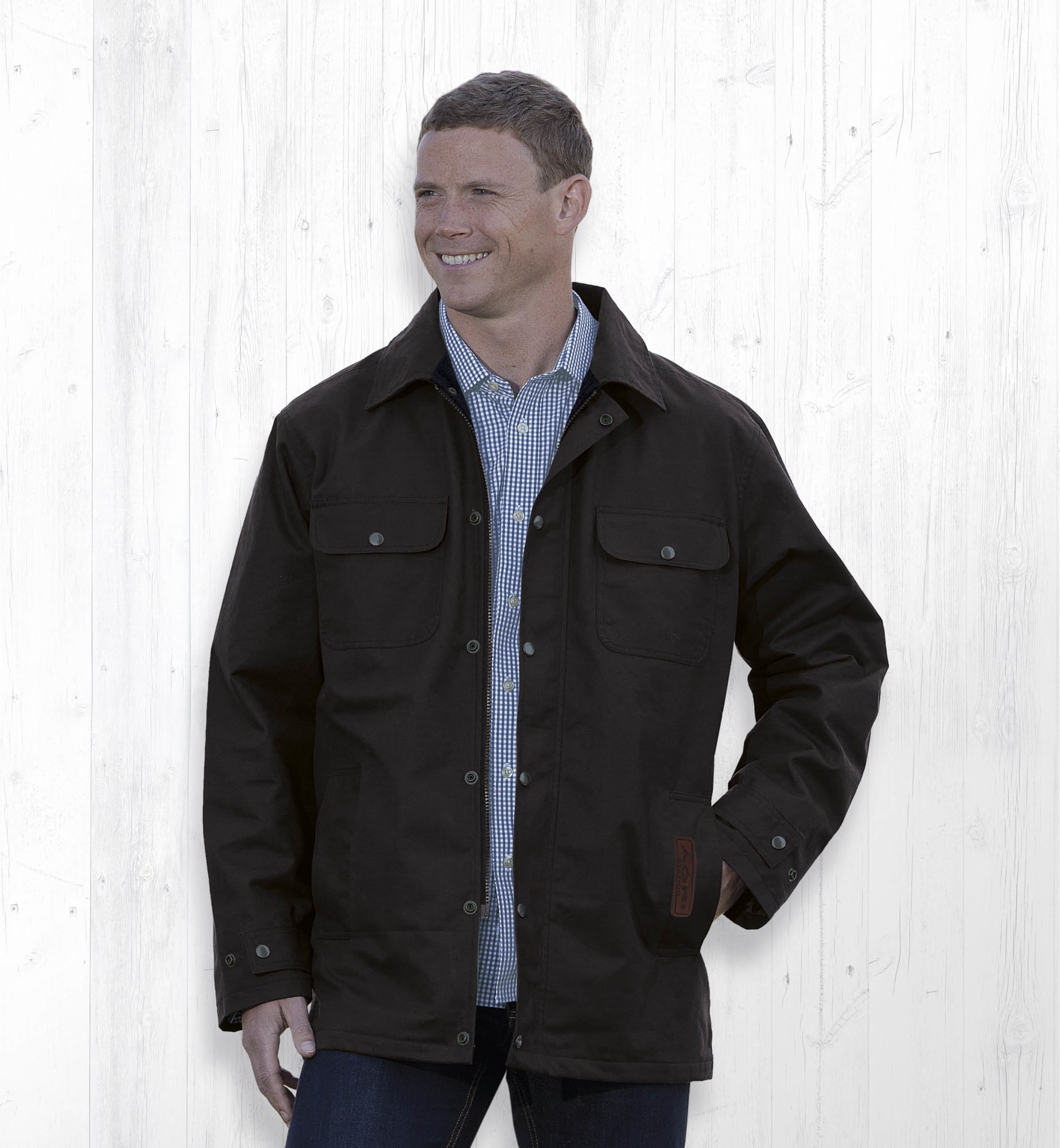 A dark brown oilskin jacket with pockets and a front button closure, displayed on a smiling model against a light background. Agri Station Tundra Oilskin Jacket - Image 1