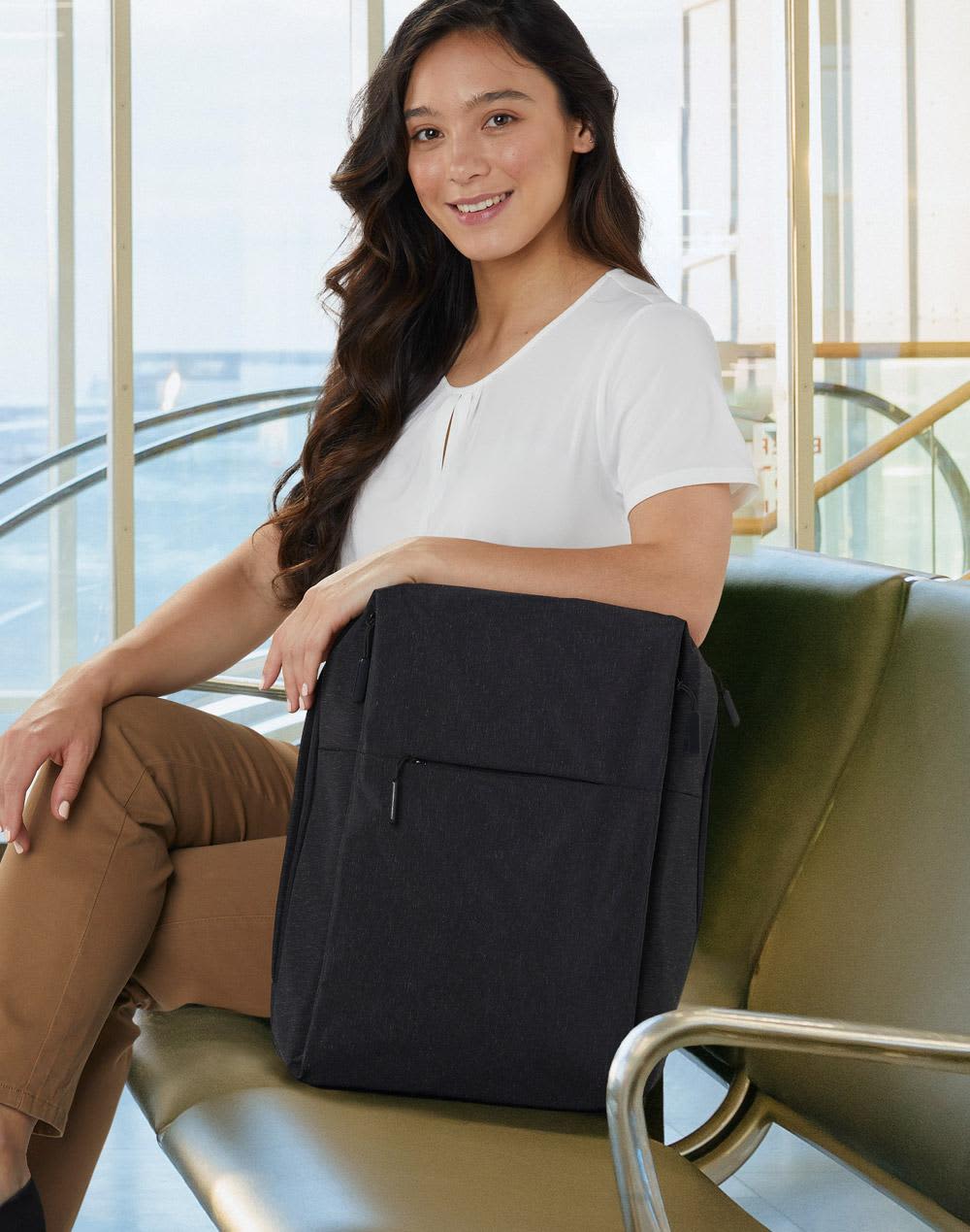 A black backpack with a front pocket, shown on a woman sitting on a green bench in a bright, modern environment. Excutive Heather Backpack - Image 1