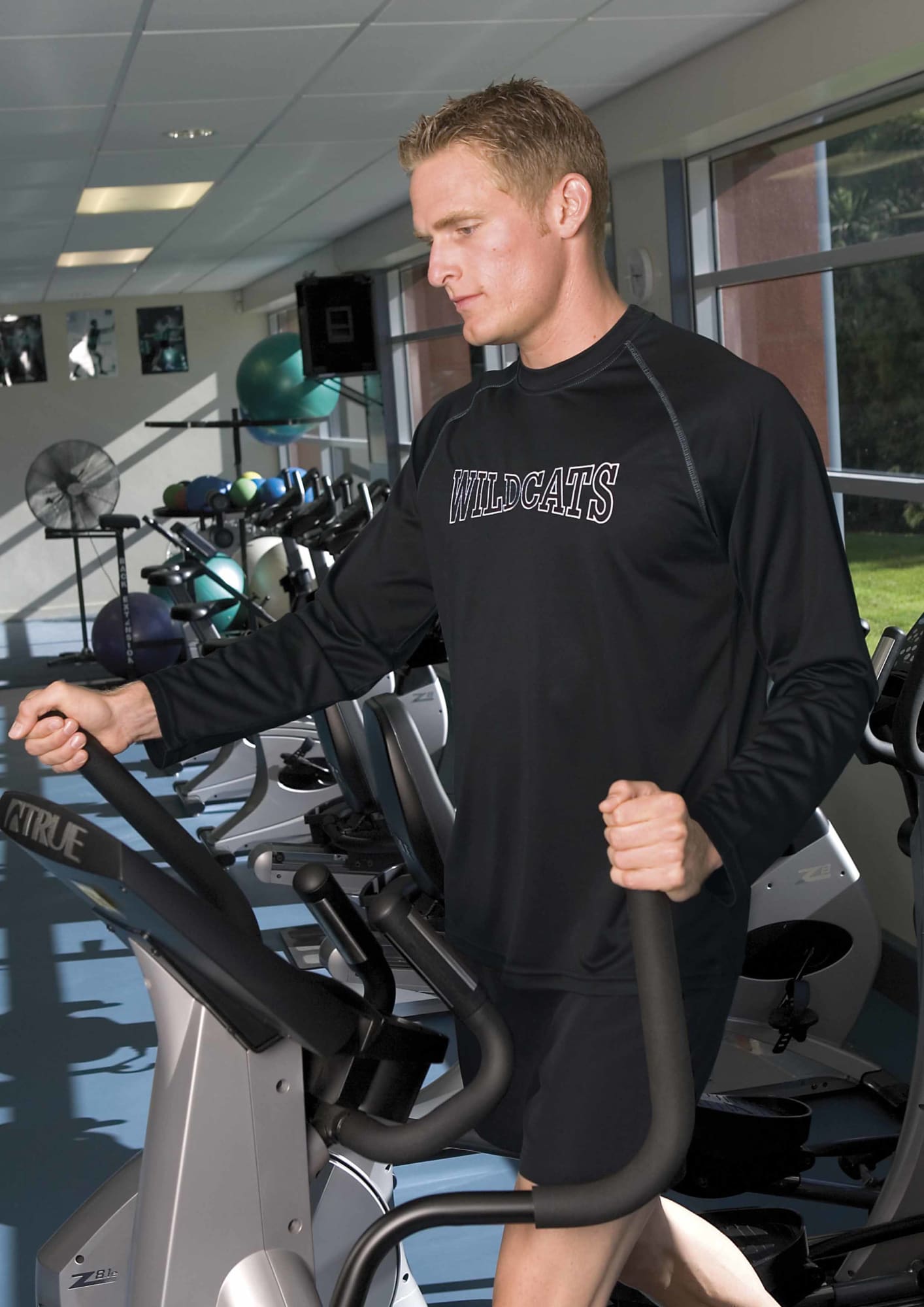 A man wearing a black long sleeve tee exercises on an elliptical machine in a gym setting. The shirt has a logo. Dri Gear Long Sleeve Tee - Mens - Image 1
