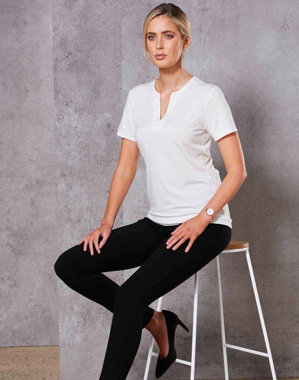A white ladies' V-neck short-sleeve knit top is worn with black pants, featuring a logo. Model seated on a bar stool. Ladies' V-neck with Tab S/S Knit Top - Image 1