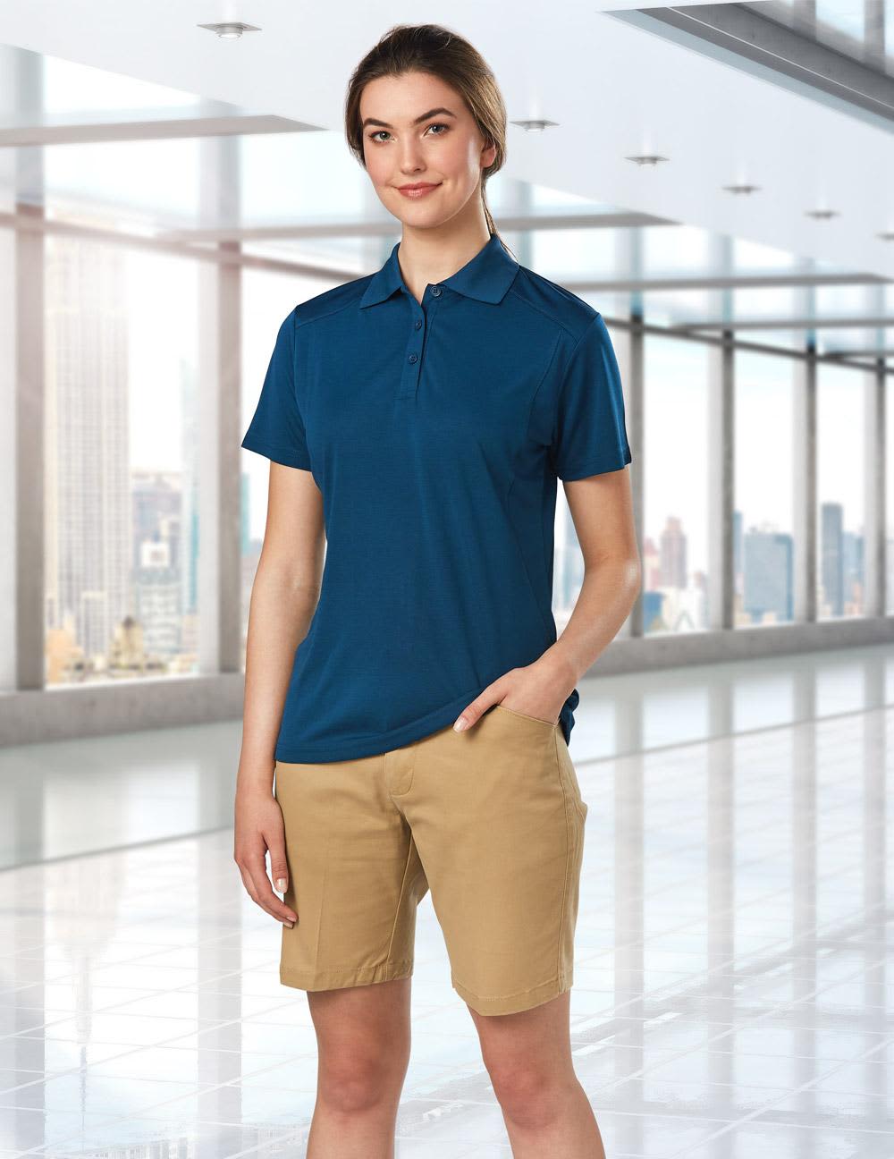 A model wears navy polo and khaki stretch cotton chino shorts in a modern indoor setting. The shorts have a logo. Ladies' Stretch Cotton Chino Shorts - Image 1