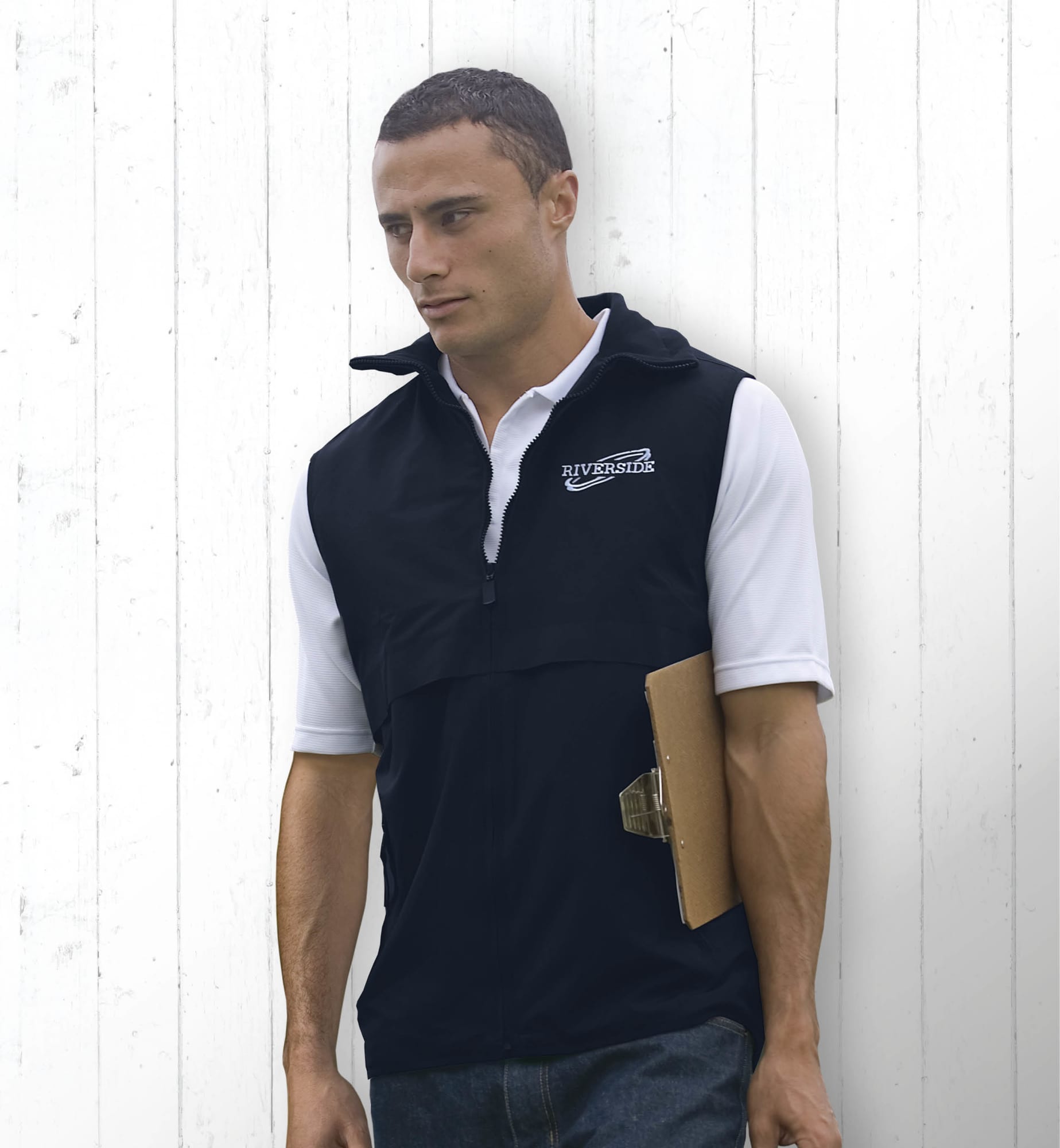 A man wearing a black nylon vest with a logo, holding a clipboard, standing against a white wooden background. Nylon Ottoman Vest - Image 1