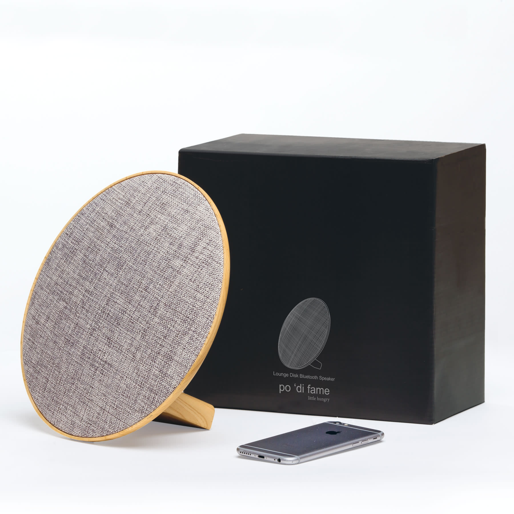 A round grey Bluetooth speaker with a wooden base next to a smartphone, accompanied by a black box packaging. Lounge Disc Bluetooth Speaker - Image 1