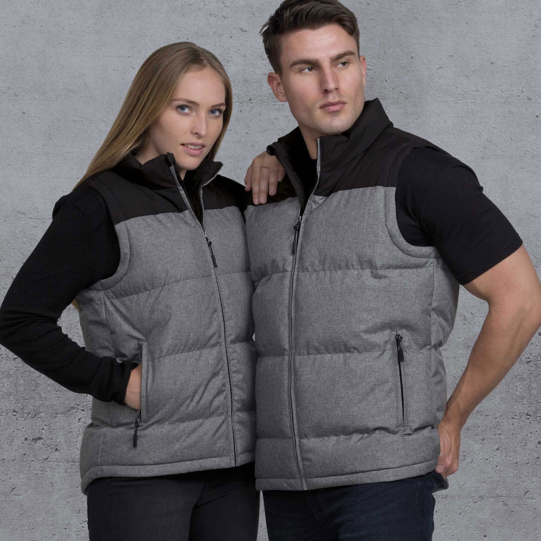 A grey and black puffa vest is worn by a man and woman against a textured background. The vest features a zip and pockets. smpli Axle Puffa Vest - Image 1