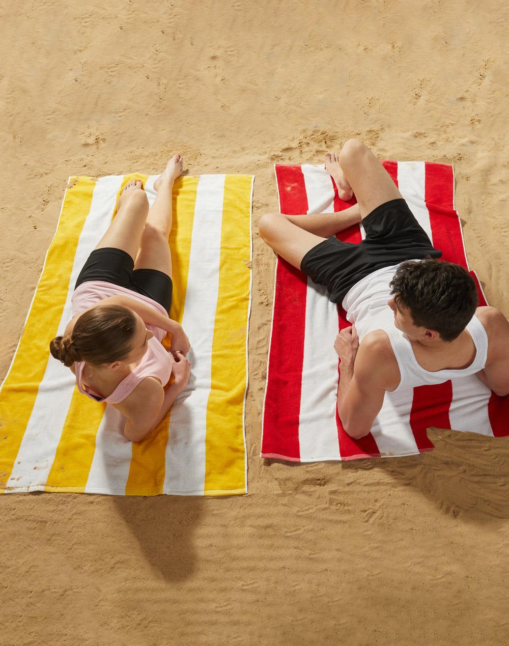 Striped beach towels in yellow and red, laid out on sand, with a man and woman relaxing on them. Striped Beach Towel - Image 1