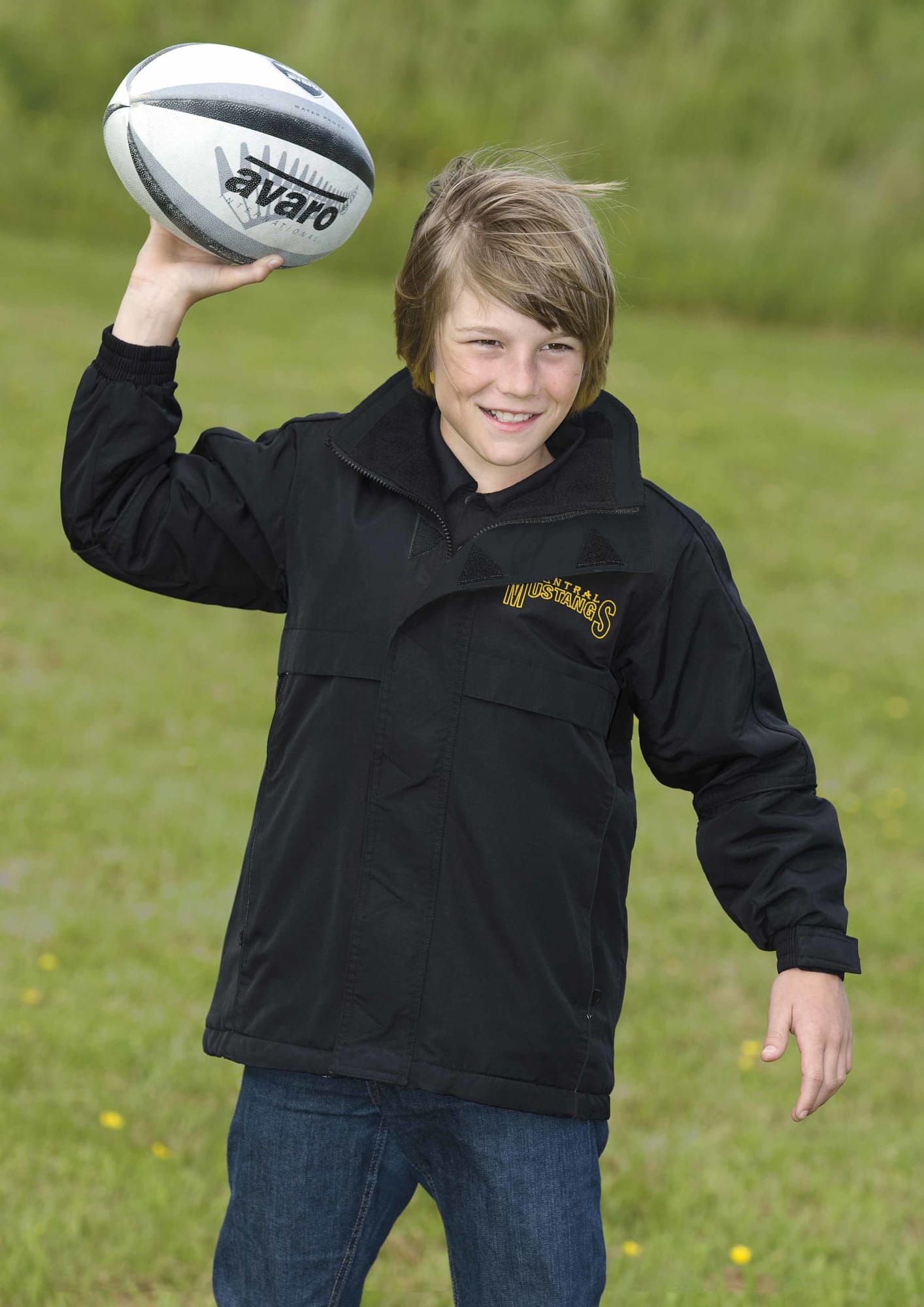 A youth wearing a black rain jacket, holding a rugby ball, standing on grass outdoors. The jacket has a logo. Youth Plain Basecamp Anorak - Image 1