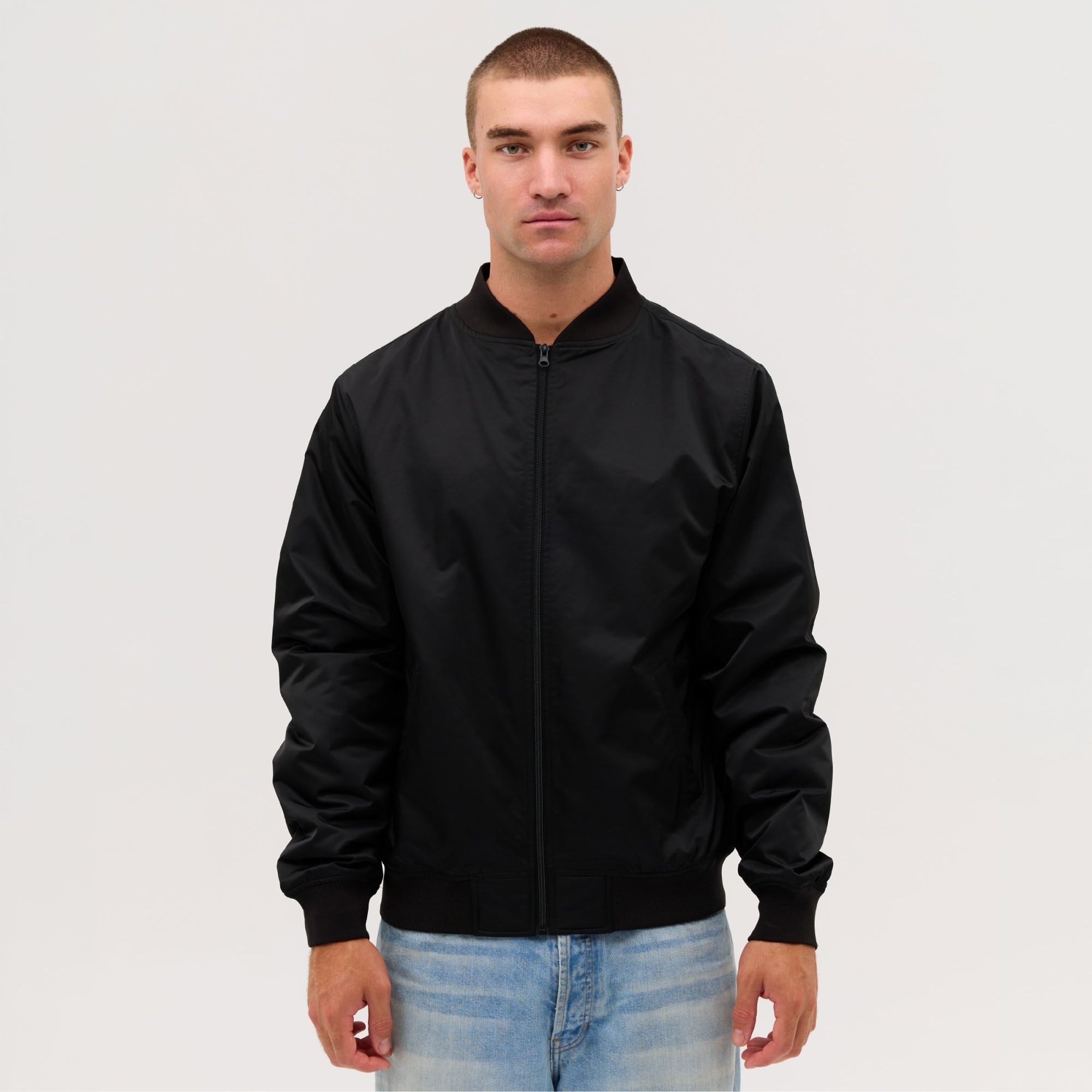 A black bomber jacket worn by a model, featuring a zip front and ribbed cuffs. The model has short hair and is standing against a neutral background. Bomber Jacket - Image 1