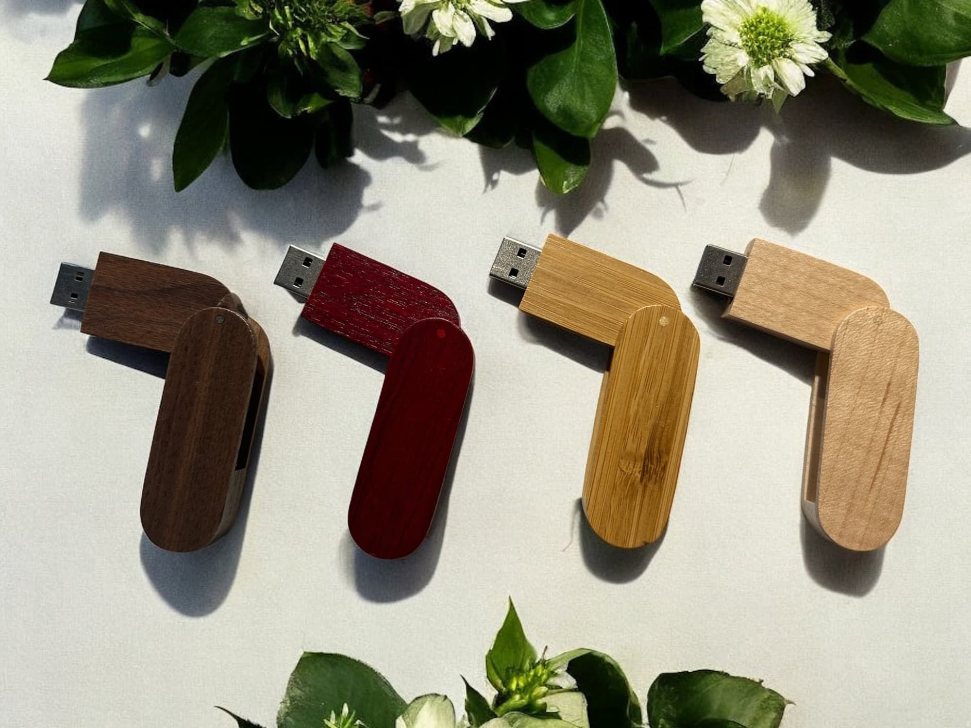 A collection of four wooden swivel flash drives in dark brown, red, light brown, and bamboo, surrounded by greenery. Driftwood Wooden Swivel 4GB Flash Drive - Image 1