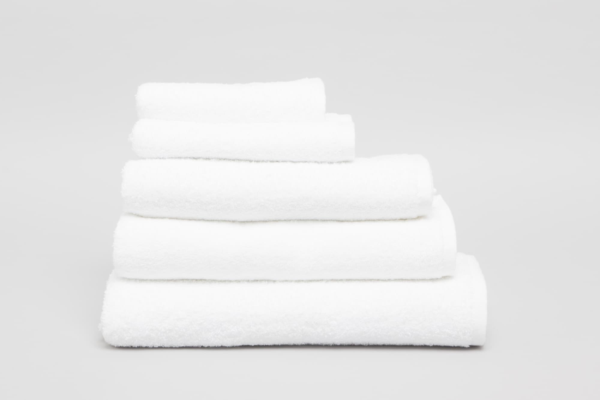 A stack of five white towels in varying sizes, showcasing their soft, fluffy texture. The image features a simple background. Elite Large Hand Towel 500 gsm White - Image 1