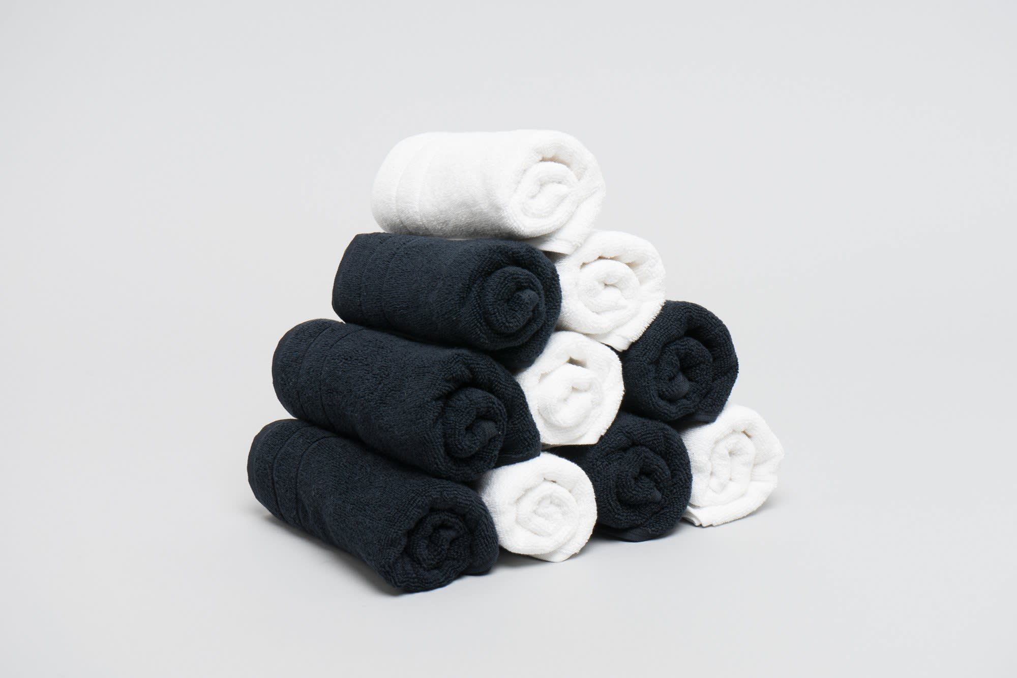 A stack of rolled towels in black and white, arranged in a pyramid shape on a light grey background. Excel Salon Towel 475 gsm White - Image 1