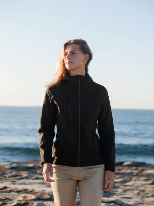 A woman stands by the beach in a black soft shell jacket, featuring a zip front and a sleek design. Kepler - Ladies - Image 1