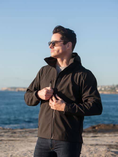 A man wearing a black soft shell jacket stands by the water, adjusting his jacket with sunglasses on. Kepler - Mens - Image 1