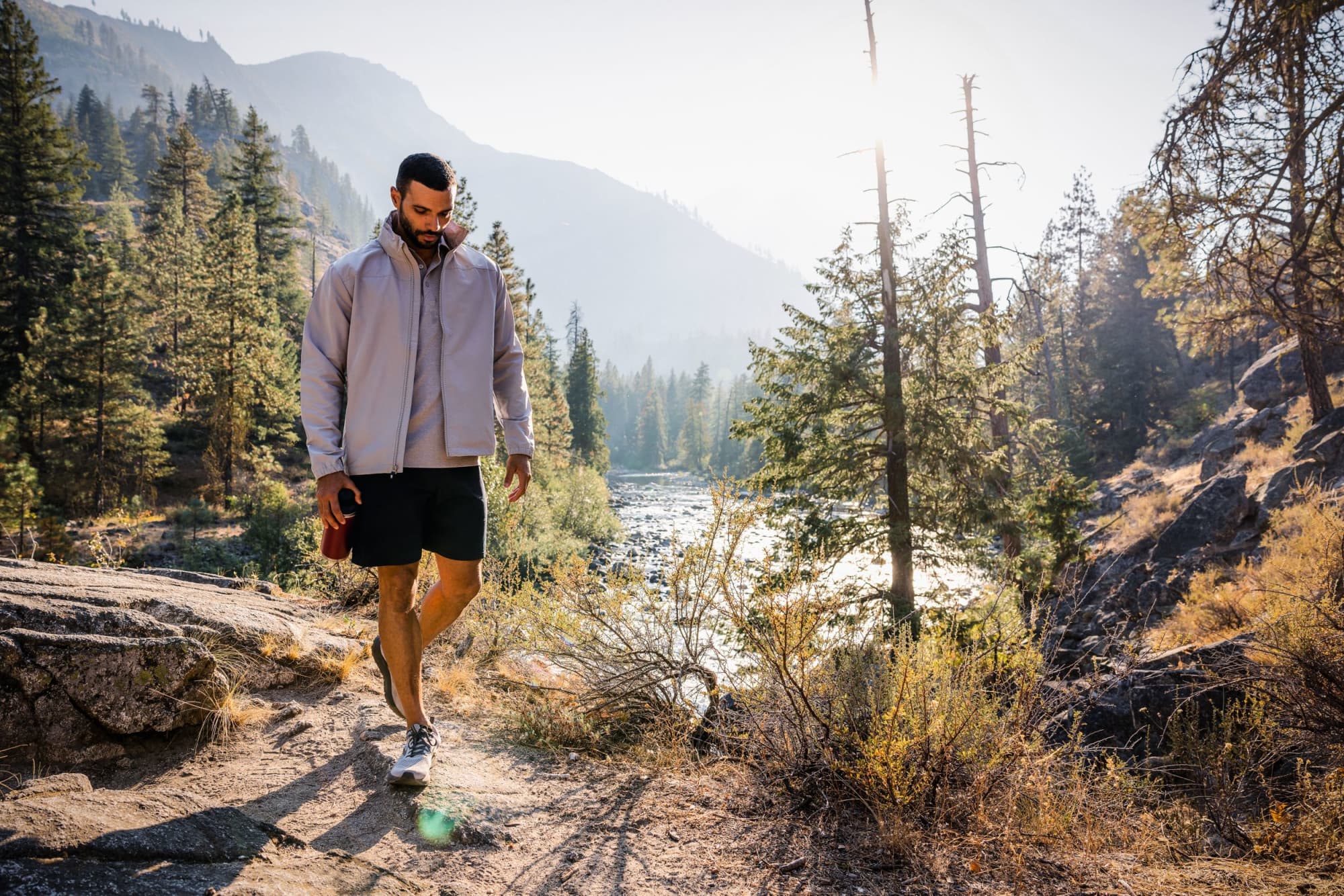 A man wearing a light grey full zip jacket and shorts walks along a rocky path near a river, surrounded by trees. Men’s Charter Full Zip Jacket - Image 1