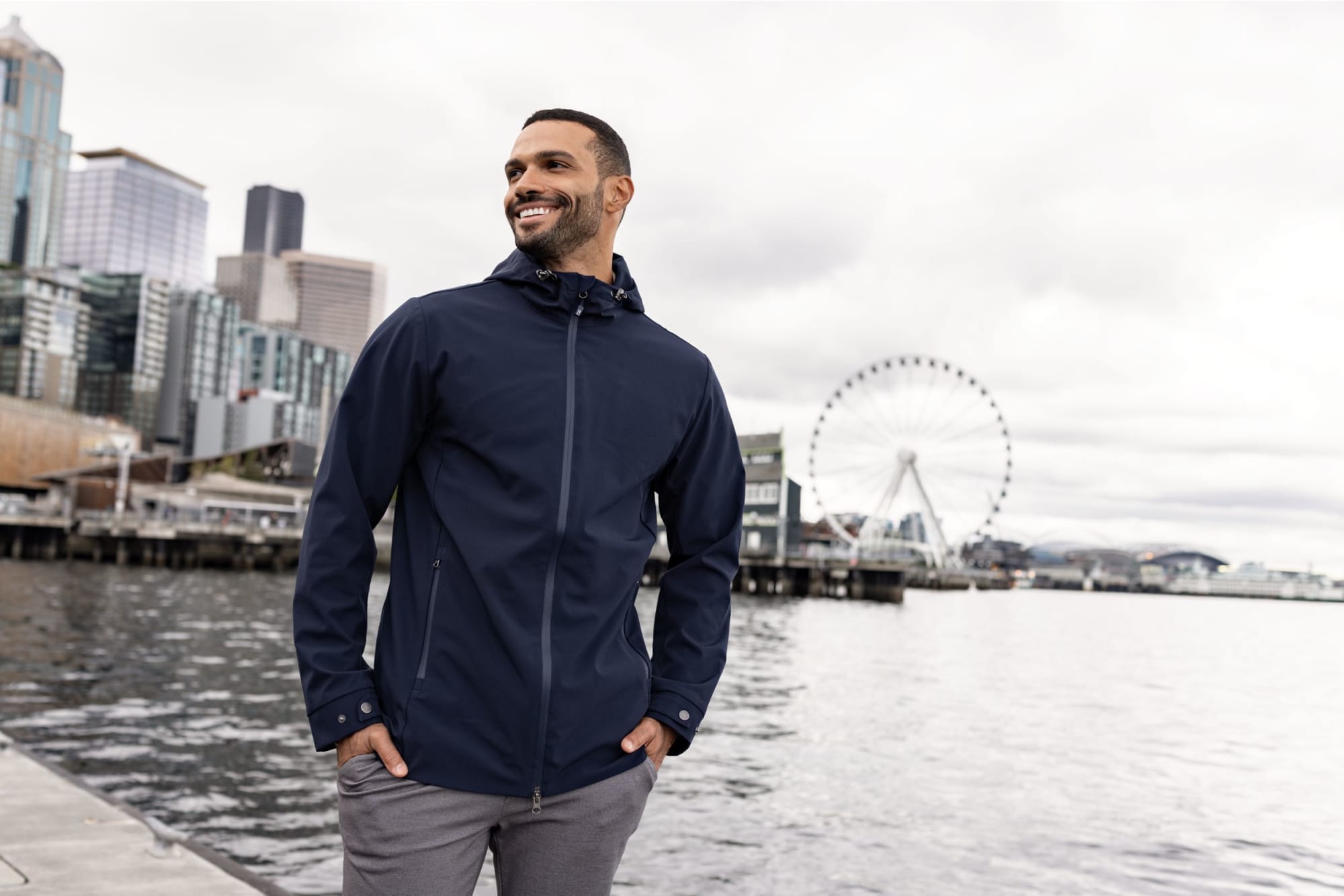 A man wearing a navy blue eco-friendly rain jacket stands by the water with a city skyline in the background. Men’s Camano Everyday Eco Jacket - Image 1