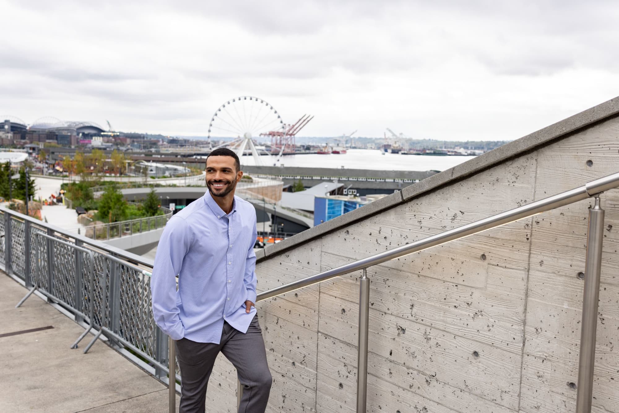 A man stands on stairs wearing a light blue pinstripe shirt and grey trousers, with a waterfront view in the background. Men's Skyline Pinstripe Shirt - Image 1
