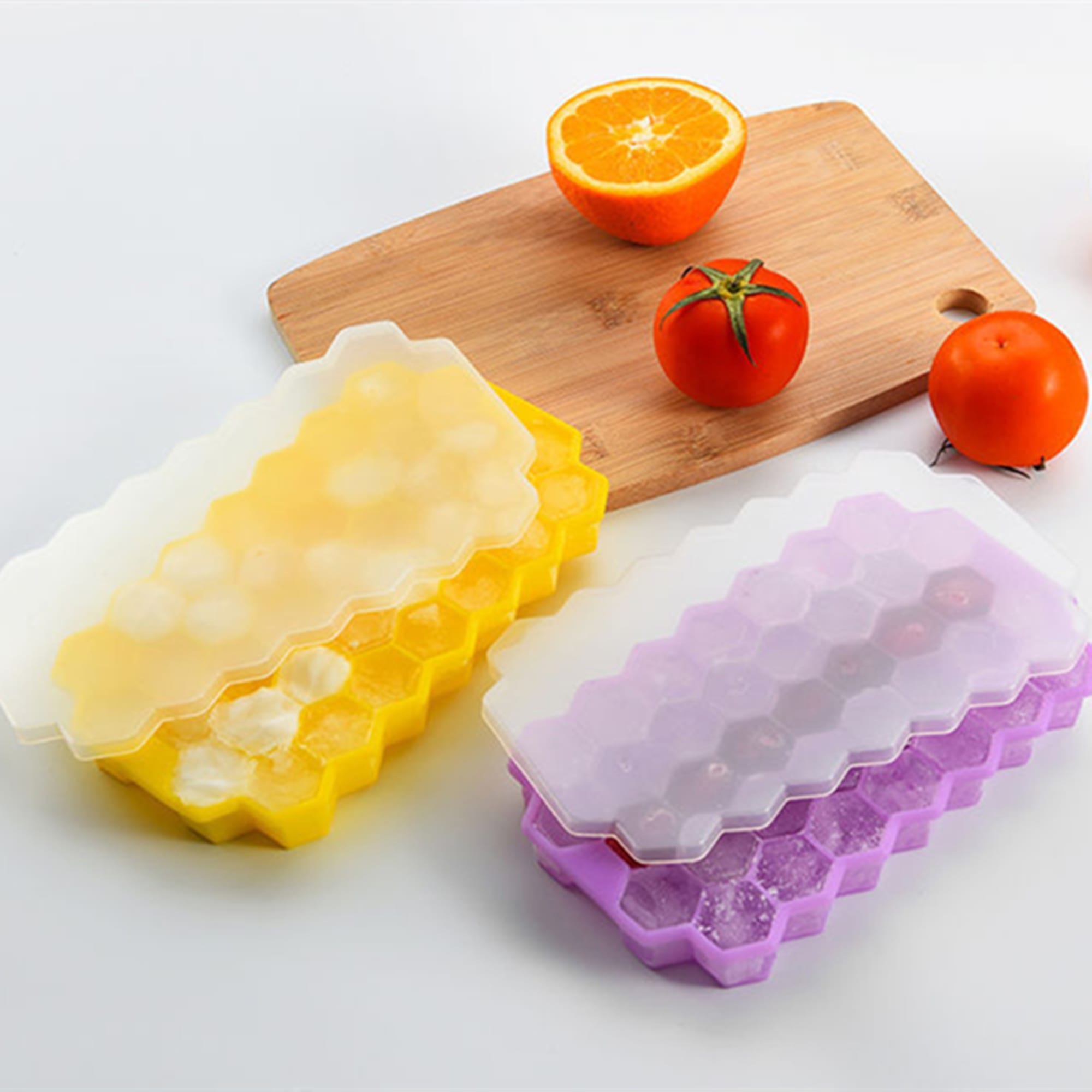 Hexagonal silicone ice cube trays in yellow and purple, with clear lids, on a wooden cutting board alongside fruits. Silicone Ice Cube Tray With Lids - Image 1