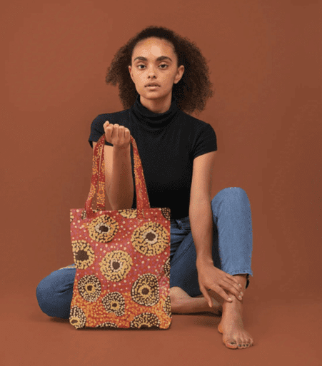 A tote bag featuring a vibrant printed design in red and yellow, held by a model against a brown backdrop. Nara RPET Sublimated Tote Bag - Image 1