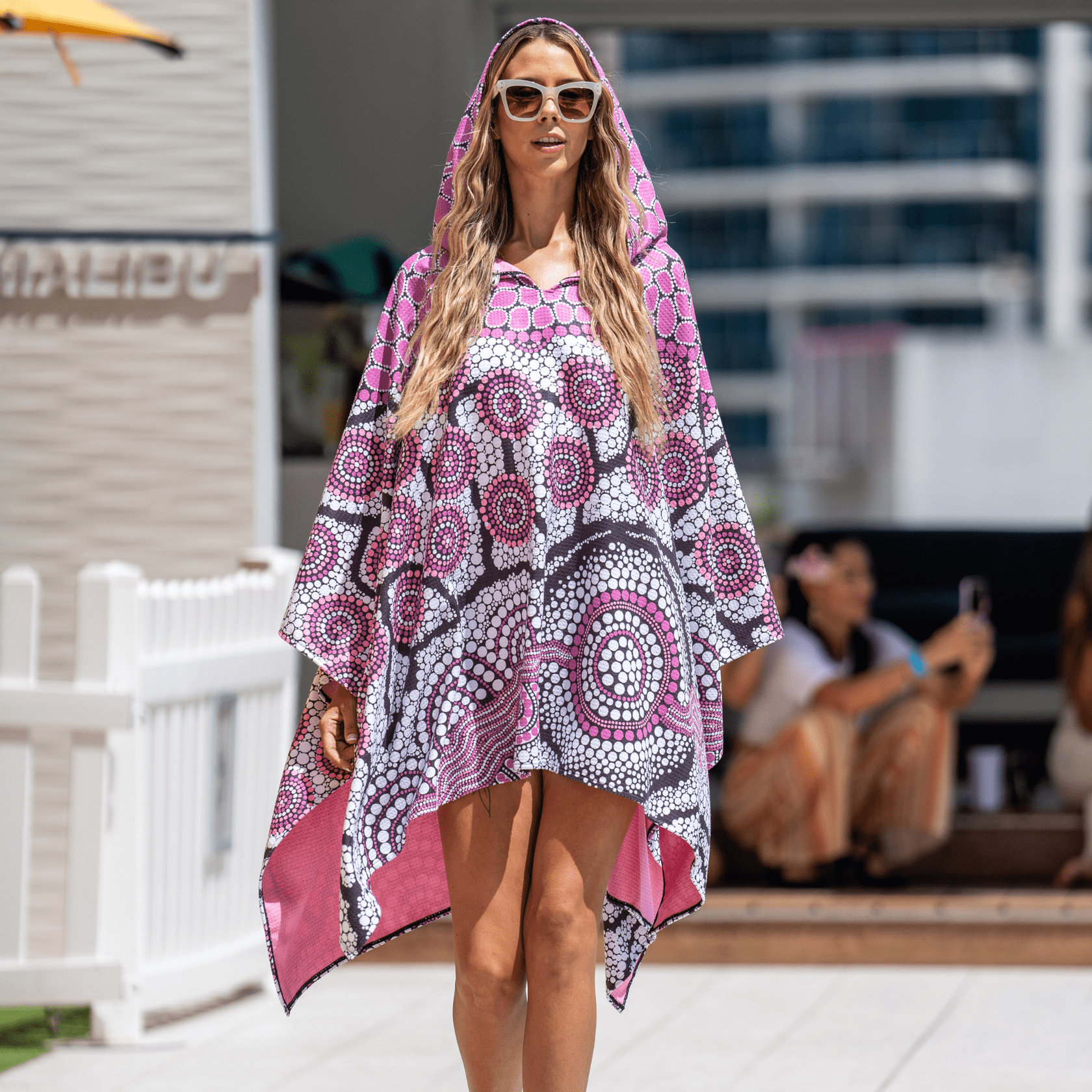 A vibrant pink and black patterned poncho with a hood, featuring circular designs. It has a logo on the front. rPET Sand Free Poncho - Image 1