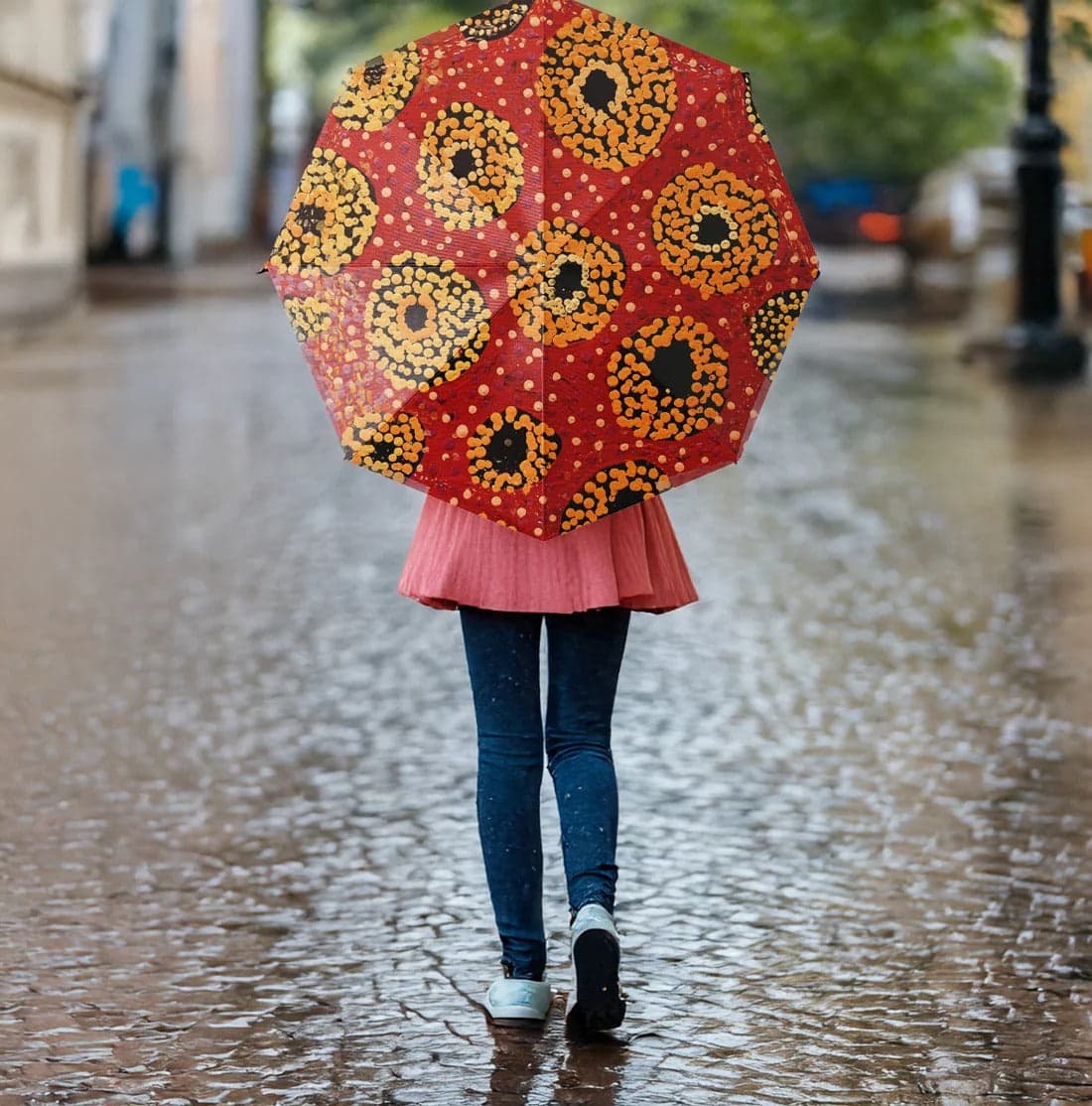 A red umbrella with a floral pattern, held by a person walking on a wet cobblestone street. Fold Away rPET Umbrella - Image 1