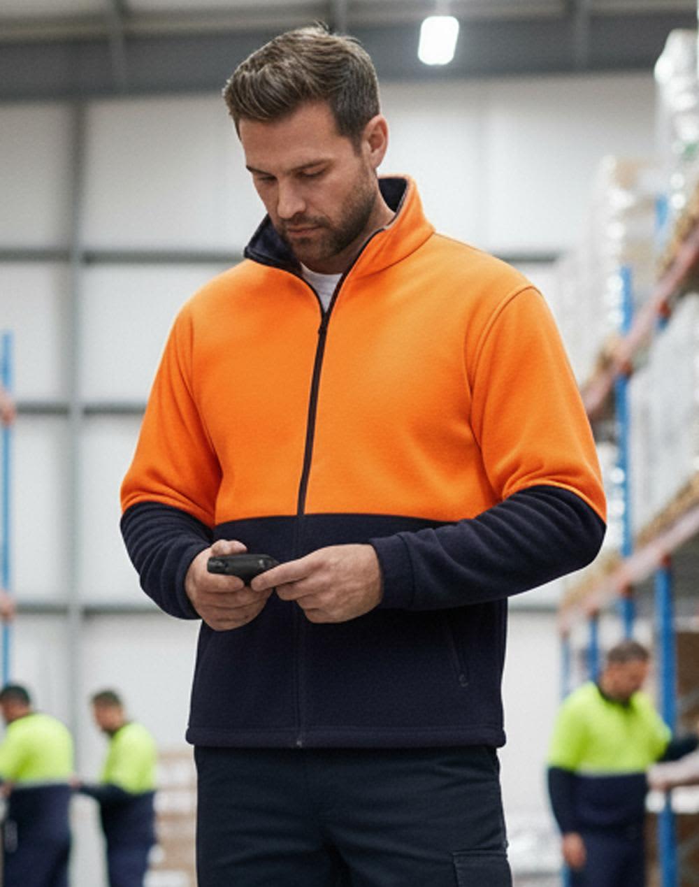 A Hi-Vis polar fleece full zip jacket in bright orange and navy, worn by a man using a phone. Hi-Vis Polar Fleece Full Zip Jacket - Image 1