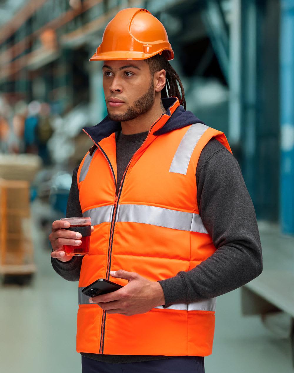 A man wearing an orange hi-vis hooded puffer vest and a hard hat stands in a warehouse, holding a coffee cup. Unisex Hi Vis Hooded Puffer Vest - Image 1