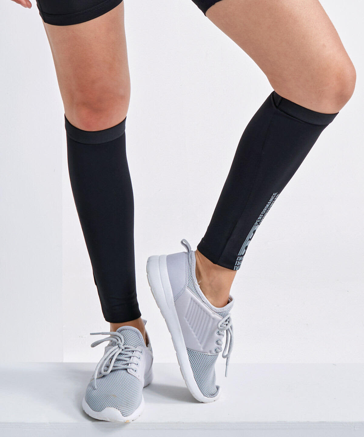 Black compression calf sleeves worn on legs, paired with light grey athletic shoes. The product features a logo. Compression Calf Sleeves - Image 1