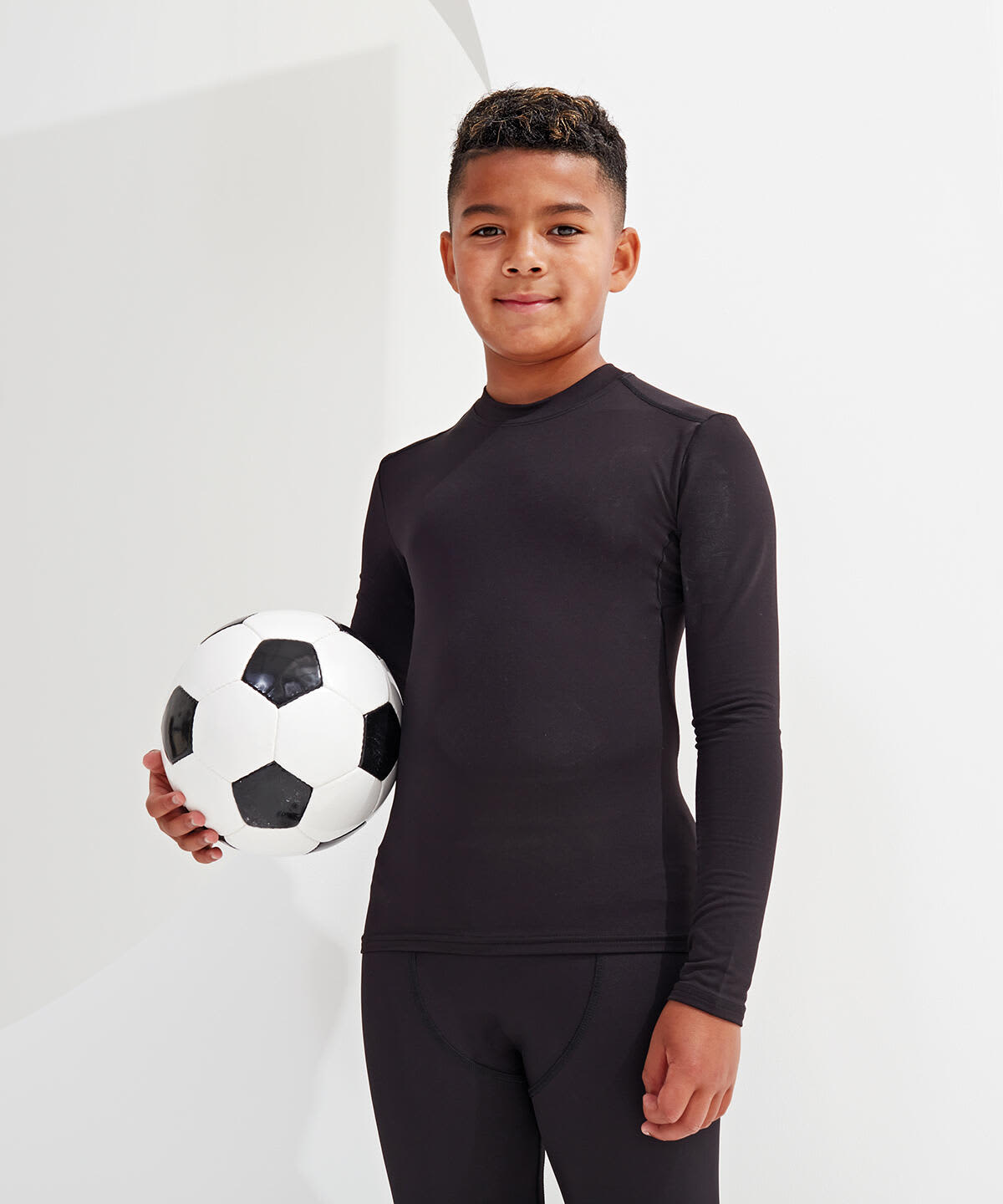 A boy stands holding a black and white soccer ball, wearing a fitted black performance baselayer with long sleeves. Kids Performance Baselayer - Image 1