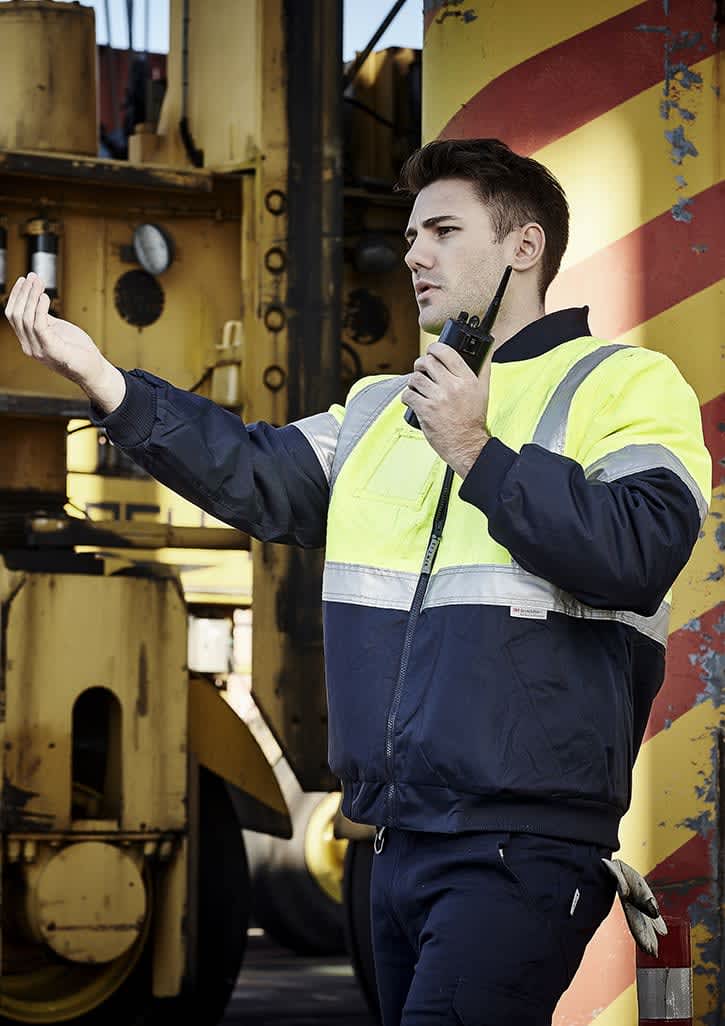 A man in a hi-vis yellow and navy quilted jacket uses a walkie-talkie against an industrial background. Mens Hi Vis Quilted Flying Jacket - Image 1