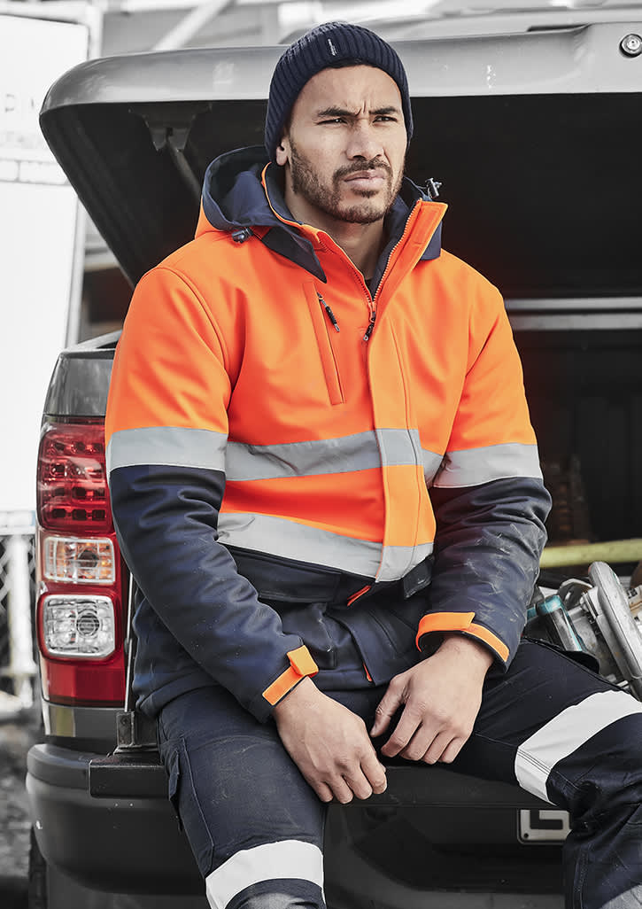 A unisex hi-vis softshell jacket in orange and navy, featuring reflective stripes, worn by a person sitting on a vehicle. Unisex Hi Vis Antarctic Softshell Jacket - Image 1