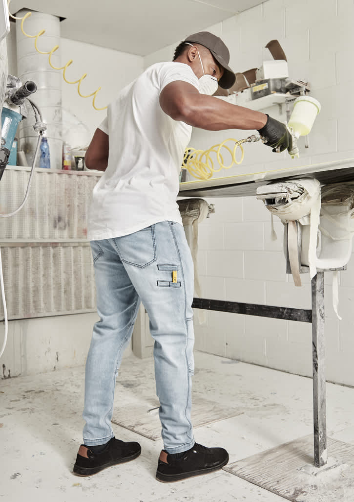 A man wearing a white shirt and grey streetwear stretch jeans, painting in a workshop setting. Mens Streetworx Stretch Jean - Image 1