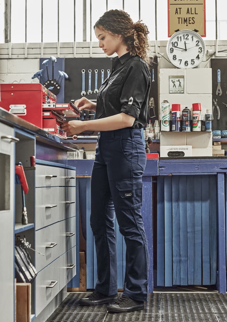 A woman in a black shirt wears navy rugged cargo pants while working in a workshop, surrounded by tools and equipment. Womens Rugged Cooling Cargo Pant - Image 1
