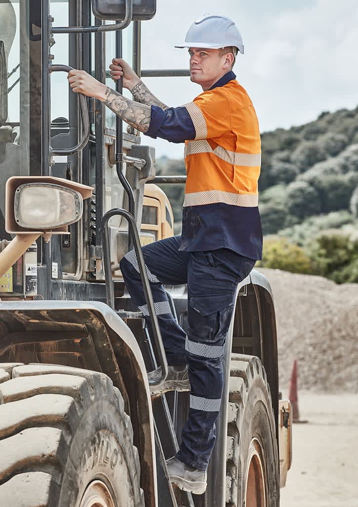 A man wearing an orange and navy shirt and dark work pants climbs into a large machinery vehicle on a worksite. Mens Rugged Cooling Stretch Taped Pant - Image 1