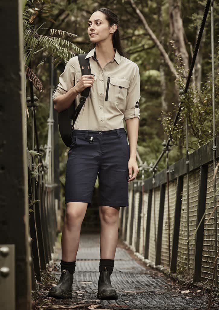 A woman stands on a wooden bridge wearing khaki and navy rugged cooling vented shorts, paired with a button-up shirt and boots. Womens Rugged Cooling Vented Short - Image 1