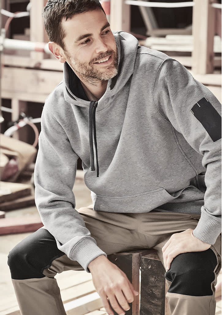 A grey unisex multi-pocket hoodie is worn by a man seated on a wooden bench, with worksite equipment in the background. Unisex Multi-Pocket Hoodie - Image 1