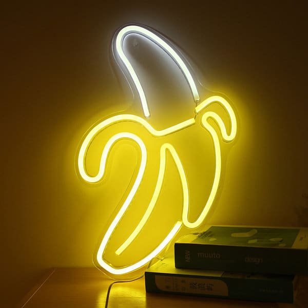 A yellow LED neon light shaped like a banana, illuminated against a wall, with books underneath. LED Neon Light - Image 1