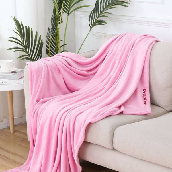 A soft pink flannel fleece blanket draped over a light-coloured sofa, with greenery in the background. Flannel Fleece Soft Blanket - Image 1