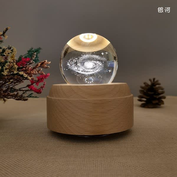 A crystal ball night light with a wooden base, featuring a galaxy design inside. Surrounding decorations include pinecones and foliage. LED Night Light Crystal Ball Music Box - Image 1