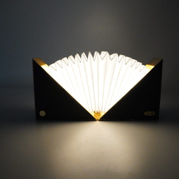 A folding book light with a black exterior and white, pleated interior, featuring gold accents. It has a logo on the side. Folding Book Light - Image 1