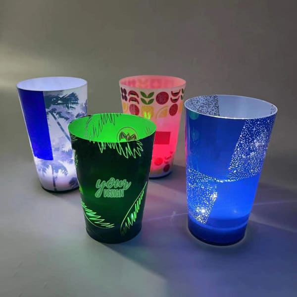 Four illuminated party cups in various designs and colours: green, blue, pink, and a patterned cup with a logo. 14 oz. LED Glowing Party Cup - Image 1