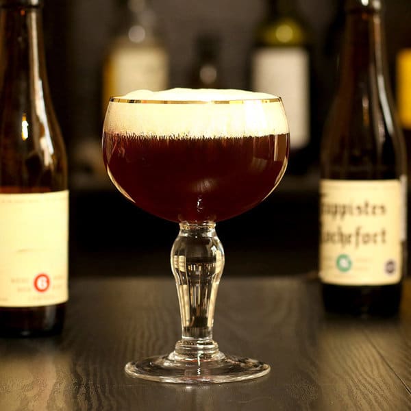 A crystal beer mug filled with dark beer topped with frothy foam, surrounded by bottles on a bar counter. Crystal Beer Mug - Image 1