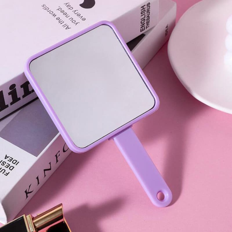 A purple square-handled mirror with a smooth reflective surface, placed on a pink background near other beauty products. 2 In1 Makeup Mirror Comb Set - Image 1