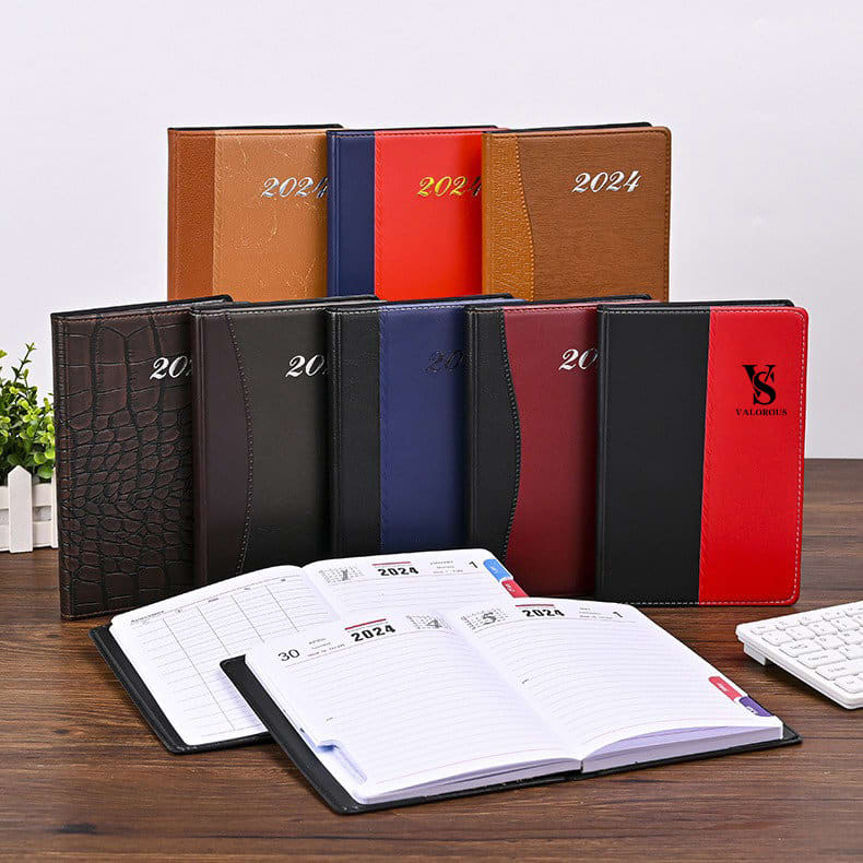 A range of PU leather diaries in various colours, including brown, red, black, and blue, displayed on a wooden table. Custom 2024 PU Leather Diary - Image 1
